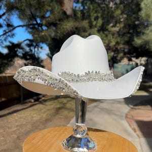 May include: A white cowboy hat with a decorative crystal band around the crown and brim. The hat is displayed on a clear, crystal-studded stand, set on a small wooden table with crystal accents. The hat's design is elegant and eye-catching.
