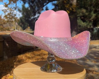 May include: A pink cowboy hat with a sparkling, glittery brim, displayed on a small wooden stool with rhinestone trim. The hat is the focal point, catching the light with its shimmering details.
