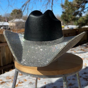 May include: A black cowboy hat with a silver rhinestone brim and accents, resting on a small wooden stool with rhinestone-covered legs. The hat is the focal point, sparkling in the sunlight.