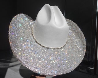 Puede incluir: Un sombrero de vaquero blanco con un ala brillante cubierta de pedrería. El sombrero presenta una corona clásica y una banda decorativa alrededor de la base de la corona. Un accesorio glamuroso.