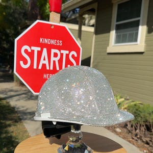 May include: A silver hard hat covered in sparkling rhinestones sits on a wooden stool with a crystal base. A red stop sign with the words "KINDNESS STARTS" is in the background.