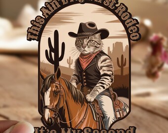 May include: A brown and white cat wearing a cowboy hat and waistcoat rides a brown horse. The cat is holding the reins and the horse is looking to the left. The text "This Ain't My First Rodeo It's My Second" is written around the image.