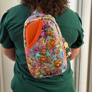 May include: A sling backpack featuring a vibrant, multi-colored pattern of orange birds, citrus fruit slices, and flowers on a purple and pink background. The bag includes a solid orange side pocket, a zipper closure, and a small orange bird-shaped charm.