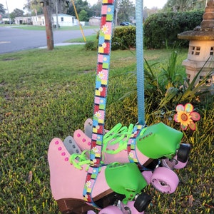 May include: Pink roller skates with green toe stops and wheels. The skates are hanging from a colorful daisy patterned strap.