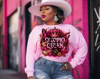 May include: A light pink sweatshirt with the text "Scorpio Queen" in a decorative font, featuring illustrations of a crown, bottles, dice, and a glass of wine. The person is wearing a white hat and blue jeans.