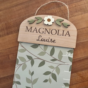 May include: A wooden growth chart featuring a light wood top plaque with the text MAGNOLIA Louise, a white flower, and green leaves. The chart has a light green background with a dark green leaf pattern and white measurement markings including 72.