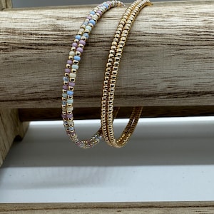 May include: Two gold beaded bracelets, one with a multi-colored pattern of pink, white, and blue beads, and the other with solid gold beads.
