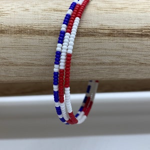 May include: A red, white, and blue beaded bracelet. The bracelet is made of small, round beads that are strung together in a pattern.