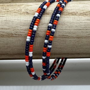 May include: Three beaded bracelets with a repeating pattern of navy blue, orange, and white beads. The bracelets are draped over a wooden dowel, showcasing the color arrangement and design. The beads are small and evenly spaced.