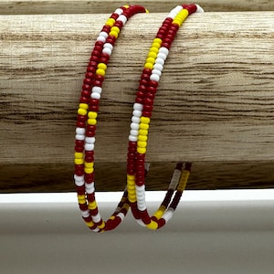 May include: Two beaded bracelets with a spiral design. The bracelets are made of small, round beads in red, yellow, and white. The bracelets are displayed on a wooden surface.
