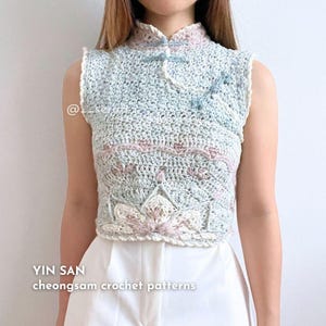 May include: A light blue crocheted cheongsam top with a high collar and a button closure. The top has a delicate floral pattern and a scalloped hem. The text "YIN SAN cheongsam crochet patterns" is visible at the bottom of the image.