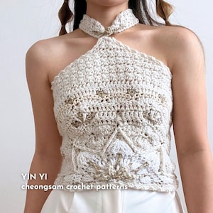 May include: A white crocheted halter top with a high neckline and a decorative gold button closure. The top features a delicate floral pattern and is styled with a white skirt. The text "YIN YI cheongsam crochet patterns" is visible at the bottom of the image.