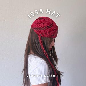 May include: A red crochet hat featuring an openwork pattern with a long, dangling tie on one side. The text ISSA HAT is printed in white above the hat, and the text crochet pattern is printed in white below the hat against a plain background.