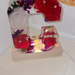 May include: A clear resin letter C with red, white, and purple flowers embedded inside. The letter is lit from within and sits on a wooden base. The text "I am Perfectly Imperfect By Ang" is visible in the background.