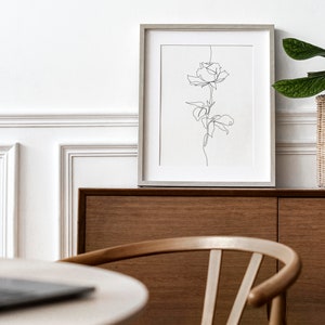 May include: A framed black and white line drawing of a rose with a wooden cabinet and a potted plant in the background.