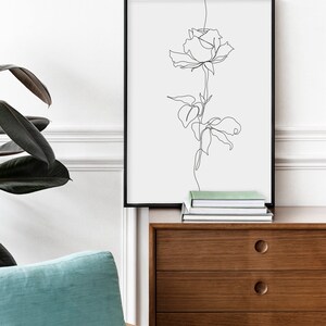 May include: A black framed print of a single line drawing of a rose with leaves on a white background. The print is hanging on a white wall above a wooden dresser with drawers.