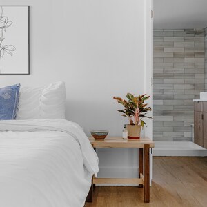 May include: A white bed with a blue pillow and white bedding. A small wooden nightstand with a plant and a bowl on top. The bedroom has white walls and a door leading to a bathroom with tiled walls.