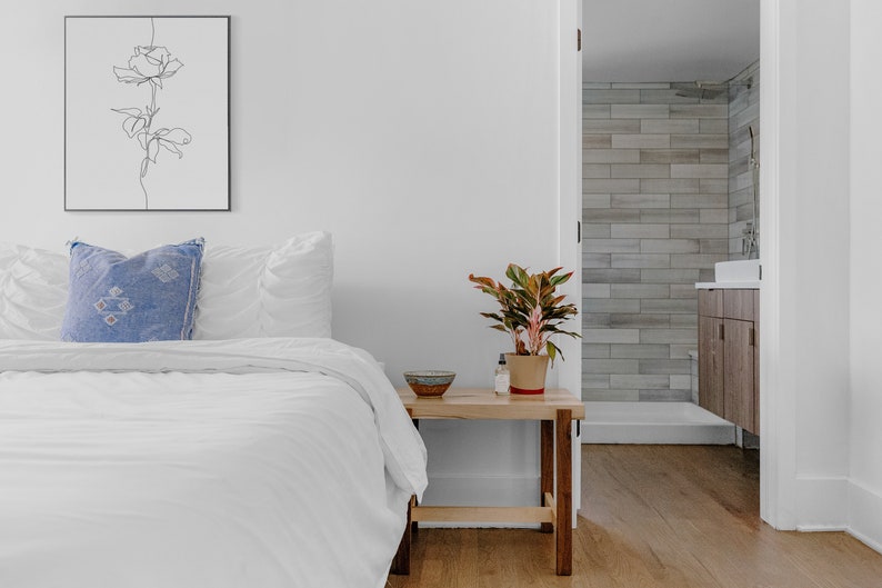 May include: A white bed with a blue pillow and white bedding. A small wooden nightstand with a plant and a bowl on top. The bedroom has white walls and a door leading to a bathroom with tiled walls.