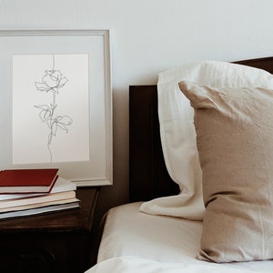 May include: A white framed print of a single line drawing of a rose on a white background. The print is on a wooden nightstand with a stack of books, including a red book on top. The print is next to a bed with white and beige bedding.