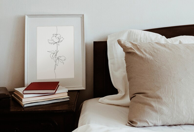May include: A white framed print of a single line drawing of a rose on a white background. The print is on a wooden nightstand with a stack of books, including a red book on top. The print is next to a bed with white and beige bedding.