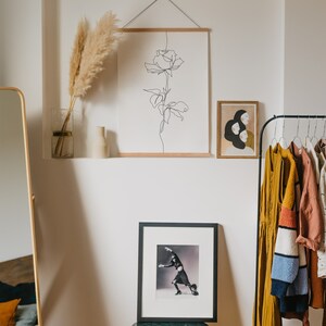 May include: A white wall with a black line drawing of a rose hanging on it. There is a framed print of two women with black hair and a rack of clothes hanging on a black metal rack. There is a small table with a black frame picture of a woman in a black dress.