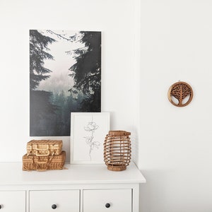 May include: A white dresser with three drawers, a wicker lantern, two wicker suitcases, a framed line drawing of a flower, and a wooden wall hanging of a tree in a circle.