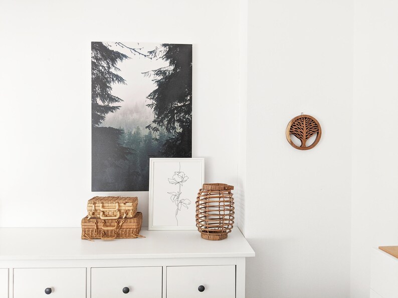 May include: A white dresser with three drawers, a wicker lantern, two wicker suitcases, a framed line drawing of a flower, and a wooden wall hanging of a tree in a circle.