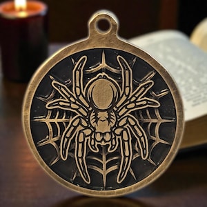May include: A round, bronze-colored pendant featuring a detailed spider design within a web. The spider is centered on a black background, with a loop at the top for hanging. The pendant has an antique, gothic aesthetic.