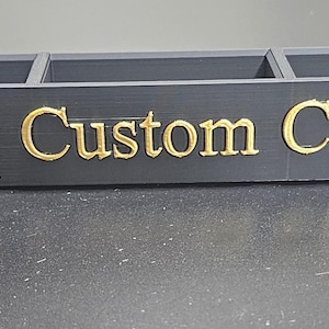 以下が含まれることがあります： 5つのコンパートメントを備えた黒いオーガナイザー。「Infinity Custom Creations」の文字が前面に金色で表示されています。オーガナイザーは長方形で、暗い面に置かれています。