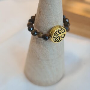 May include: A beaded ring with brown and gold beads and a gold-colored oval centerpiece. The centerpiece features a tree of life design. The ring is displayed on a light-colored wooden cone.