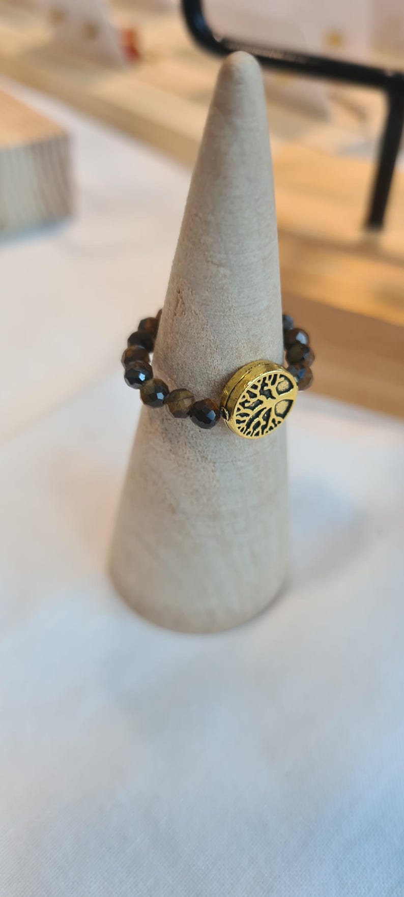 May include: A beaded ring with brown and gold beads and a gold-colored oval centerpiece. The centerpiece features a tree of life design. The ring is displayed on a light-colored wooden cone.