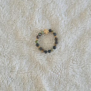 Puede incluir: Un anillo de cuentas con un dije de tr&eacute;bol de cuatro hojas de color dorado. El anillo presenta una mezcla de cuentas verdes oscuras, marrones y negras, con espaciadores dorados. El anillo est&aacute; sobre un fondo blanco texturizado.