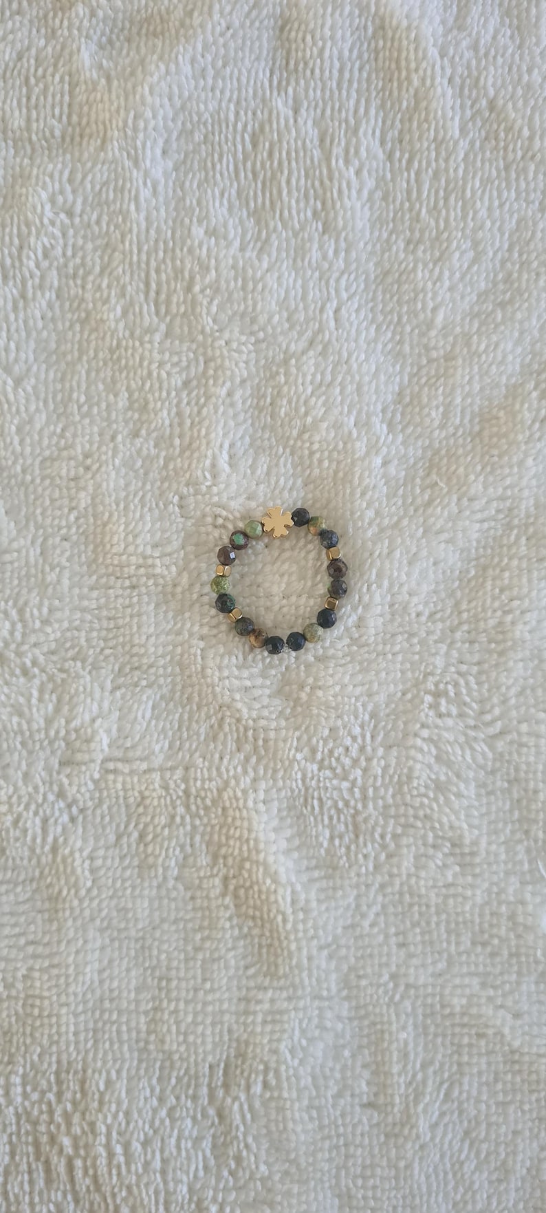 Puede incluir: Un anillo de cuentas con un dije de tr&eacute;bol de cuatro hojas de color dorado. El anillo presenta una mezcla de cuentas verdes oscuras, marrones y negras, con espaciadores dorados. El anillo est&aacute; sobre un fondo blanco texturizado.
