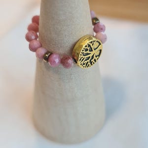 May include: A beaded ring with pink gemstones and a gold-colored accent featuring a tree design. The ring is displayed on a light-colored, cone-shaped stand. The gemstones are a soft pink color.