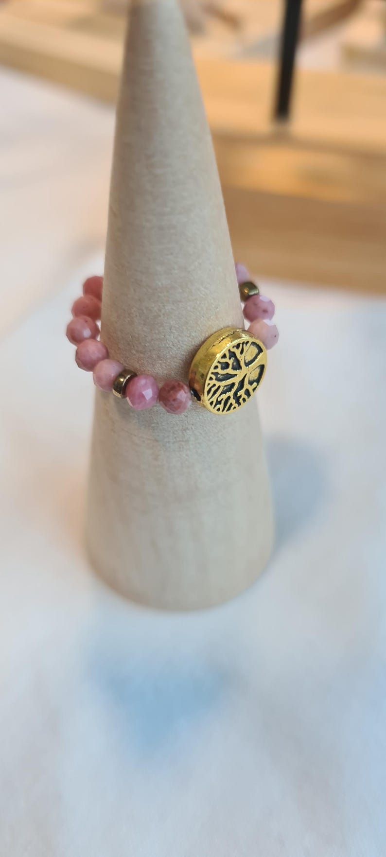 May include: A beaded ring with pink gemstones and a gold-colored accent featuring a tree design. The ring is displayed on a light-colored, cone-shaped stand. The gemstones are a soft pink color.