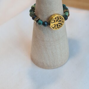 May include: A gold-colored ring with a tree of life design. The ring features a band of small, round, green and black beads. The ring is displayed on a light brown, cone-shaped stand.