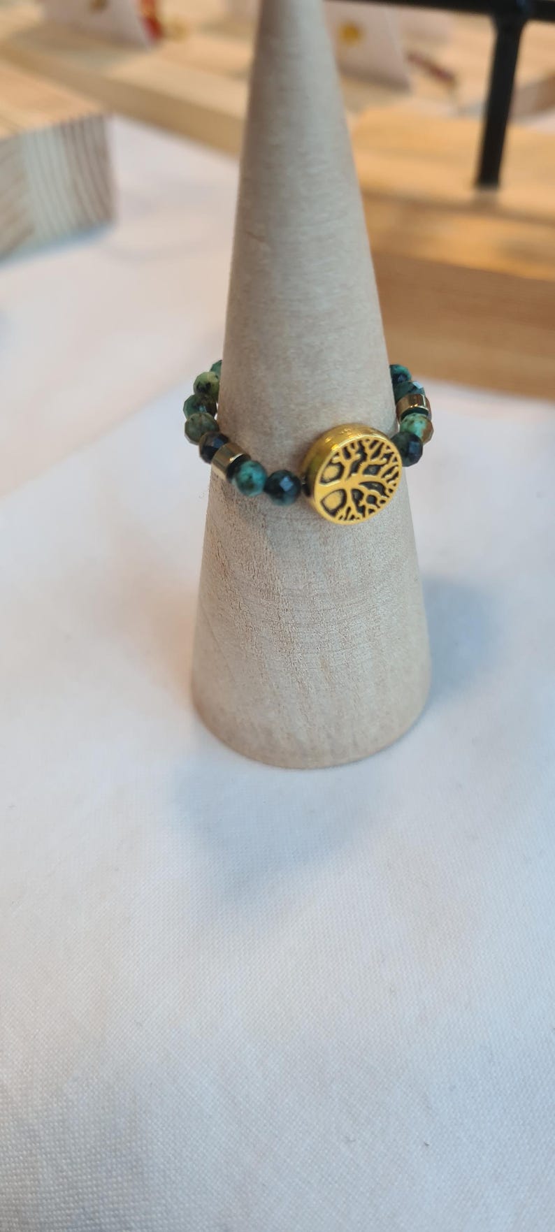 May include: A gold-colored ring with a tree of life design. The ring features a band of small, round, green and black beads. The ring is displayed on a light brown, cone-shaped stand.