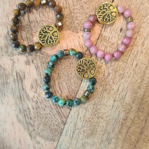 May include: Three beaded bracelets with a gold-colored tree of life charm. One bracelet is brown, one is pink, and one is teal. The bracelets are on a wooden surface.
