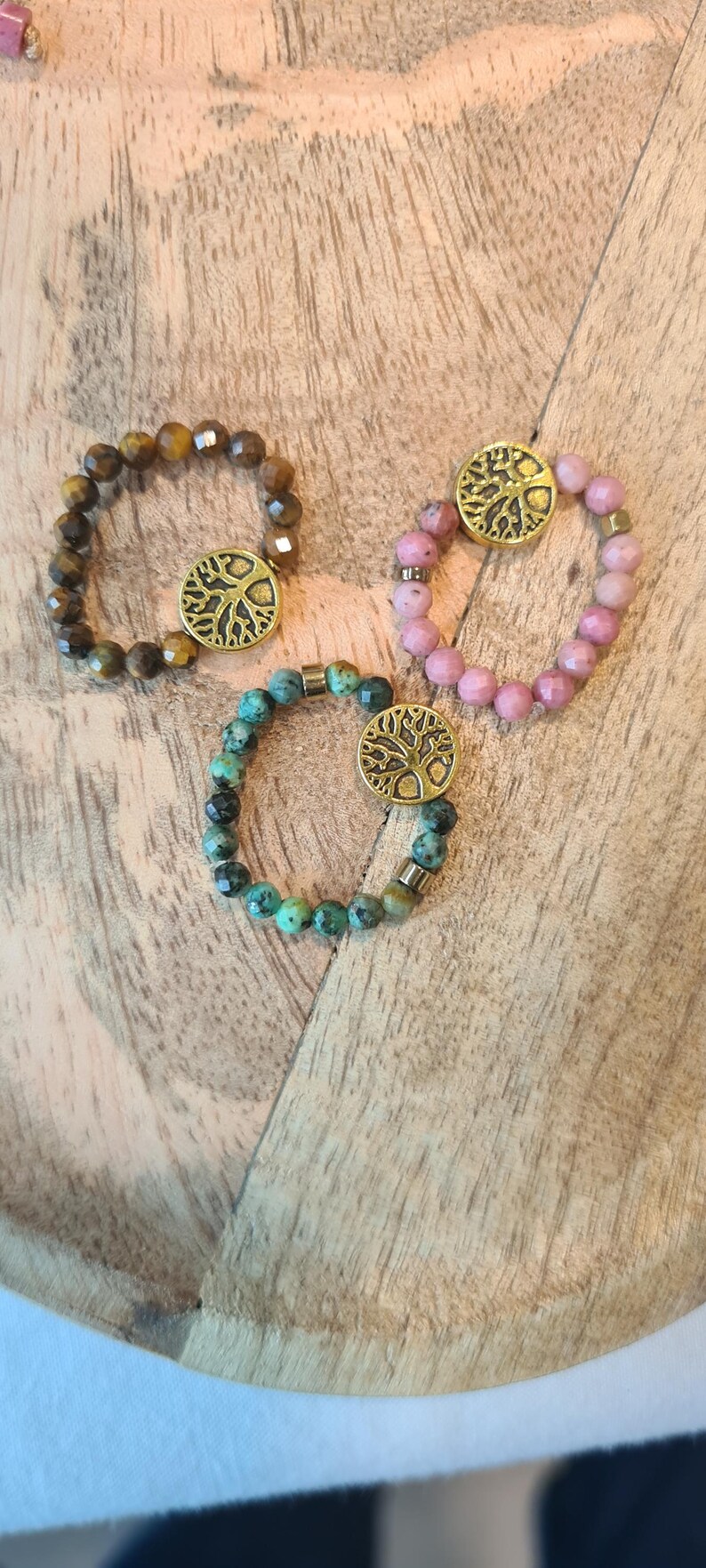 May include: Three beaded bracelets with a gold-colored tree of life charm. One bracelet is brown, one is pink, and one is teal. The bracelets are on a wooden surface.