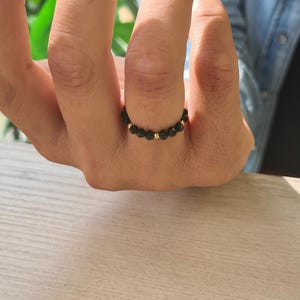May include: A black beaded ring with gold-coloured accents. The ring is made of small, round black beads and interspersed with gold-coloured beads. The ring is on a finger.