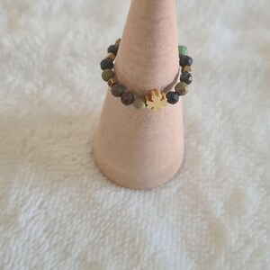 Puede incluir: Un anillo de cuentas con un dije de cruz dorada. El anillo presenta una mezcla de cuentas verdes, marrones y negras. El anillo se exhibe sobre un soporte en forma de cono marr&oacute;n claro sobre un fondo blanco texturizado.