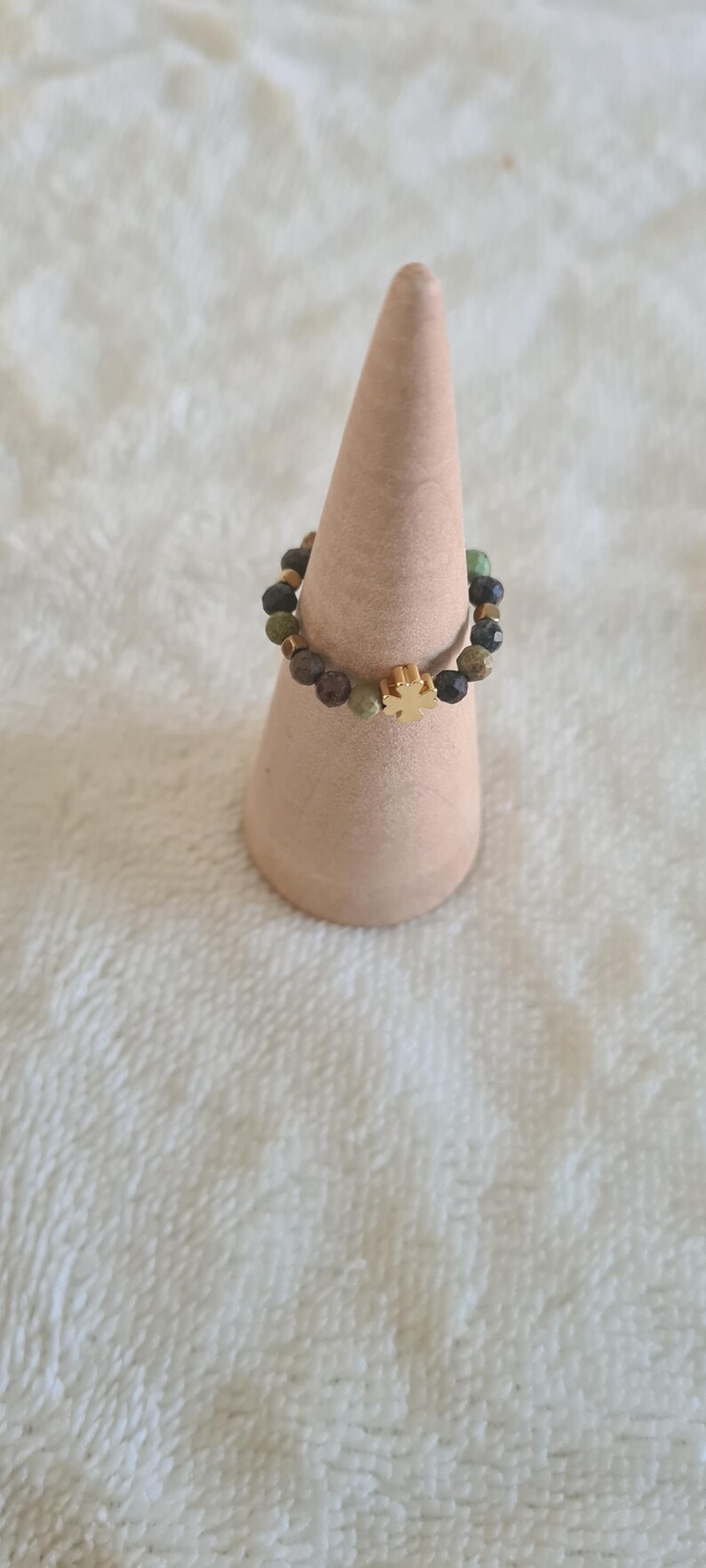 Puede incluir: Un anillo de cuentas con un dije de cruz dorada. El anillo presenta una mezcla de cuentas verdes, marrones y negras. El anillo se exhibe sobre un soporte en forma de cono marr&oacute;n claro sobre un fondo blanco texturizado.