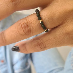 May include: A beaded ring with dark green beads and gold-coloured accents. The ring is worn on a finger, highlighting its design and colour contrast. The ring is a piece of jewellery.