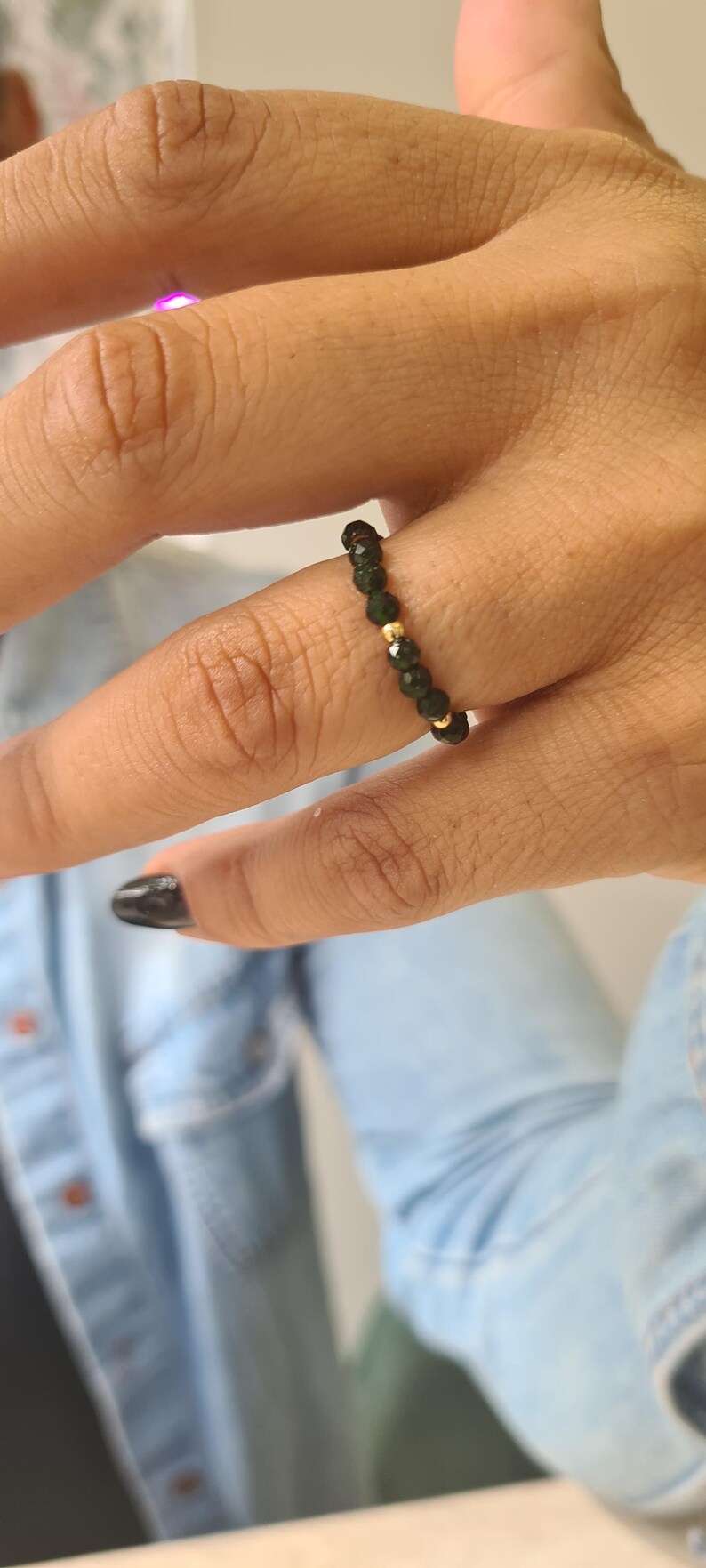 May include: A beaded ring with dark green beads and gold-coloured accents. The ring is worn on a finger, highlighting its design and colour contrast. The ring is a piece of jewellery.
