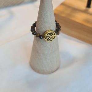 May include: A beaded ring with a gold-colored tree of life charm. The ring features small, brown beads and a circular charm with a detailed tree design. The ring is displayed on a light-colored, cone-shaped stand.