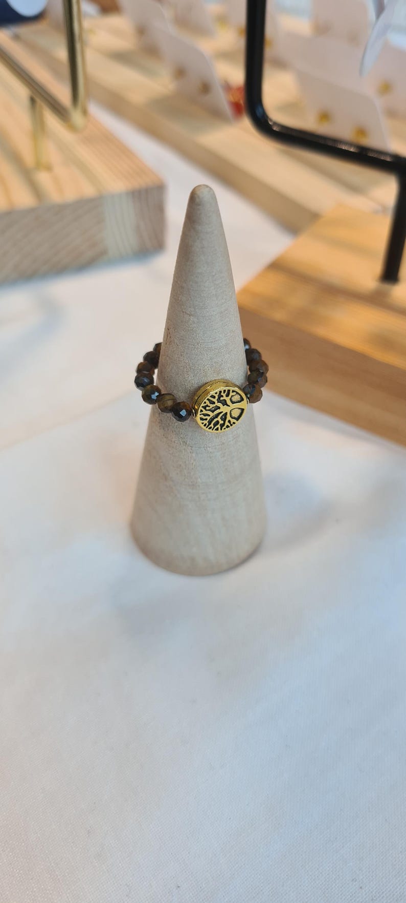 May include: A beaded ring with a gold-colored tree of life charm. The ring features small, brown beads and a circular charm with a detailed tree design. The ring is displayed on a light-colored, cone-shaped stand.
