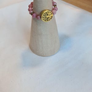 May include: A beaded ring with pink gemstones and a gold-colored tree of life emblem. The ring is displayed on a light brown cone-shaped stand, showcasing the jewelry's design and craftsmanship.