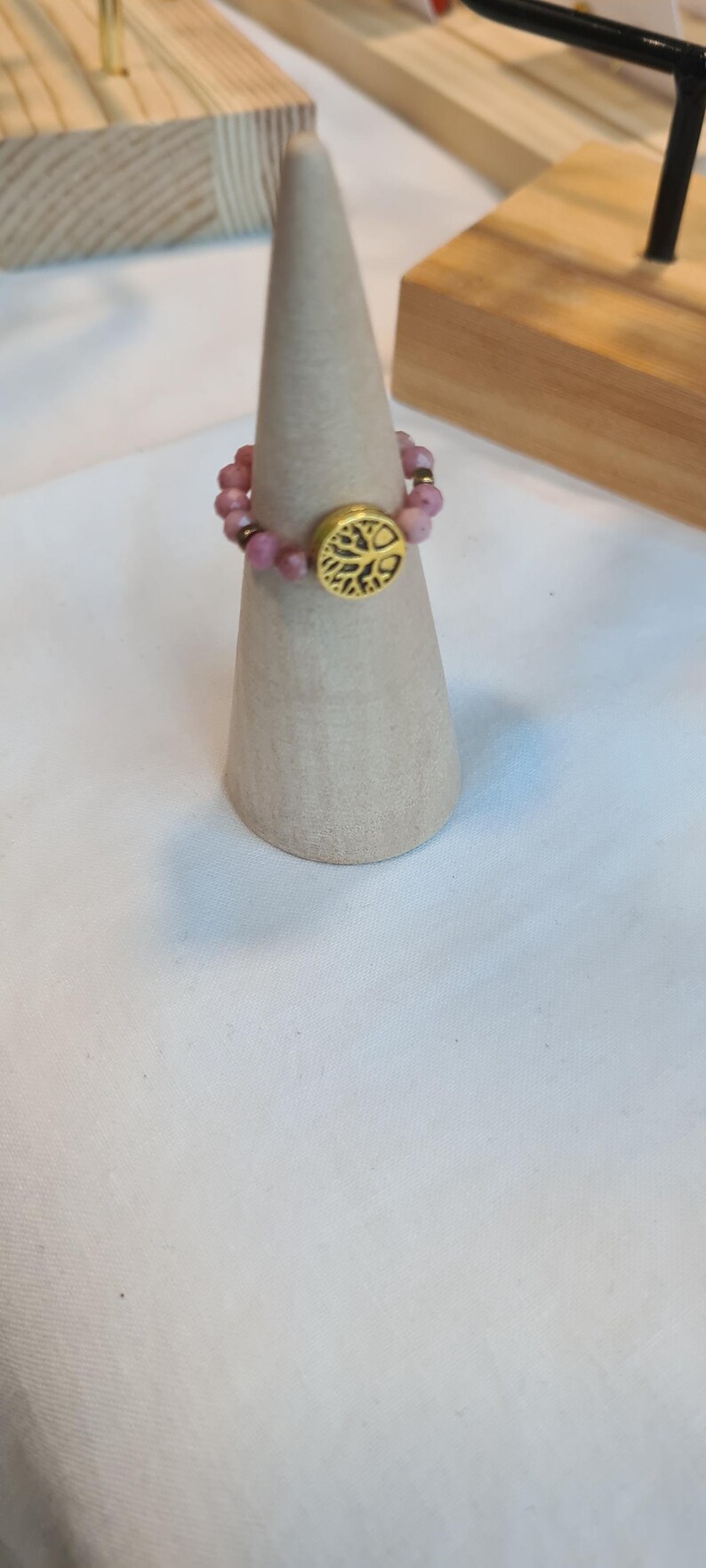 May include: A beaded ring with pink gemstones and a gold-colored tree of life emblem. The ring is displayed on a light brown cone-shaped stand, showcasing the jewelry's design and craftsmanship.