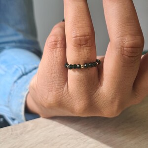 May include: A black beaded ring with gold accents is worn on a finger. The ring is made of small, dark beads and gold-coloured beads. The person's nails are painted black. The background is a light-coloured wooden surface.