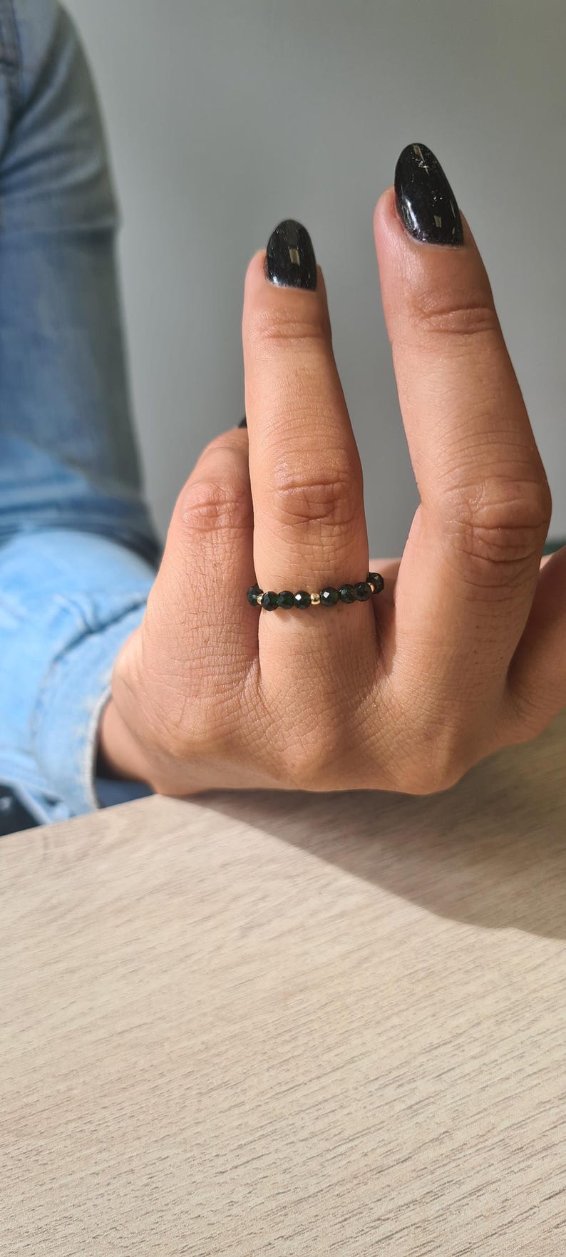 May include: A black beaded ring with gold accents is worn on a finger. The ring is made of small, dark beads and gold-coloured beads. The person's nails are painted black. The background is a light-coloured wooden surface.